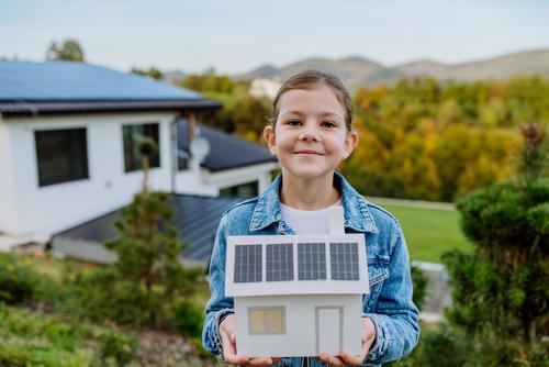 Mädchen hält Modellhaus mit Photovoltaik auf dem Dach in den Händen und steht vor einem Haus mit Photovoltaikanlage.