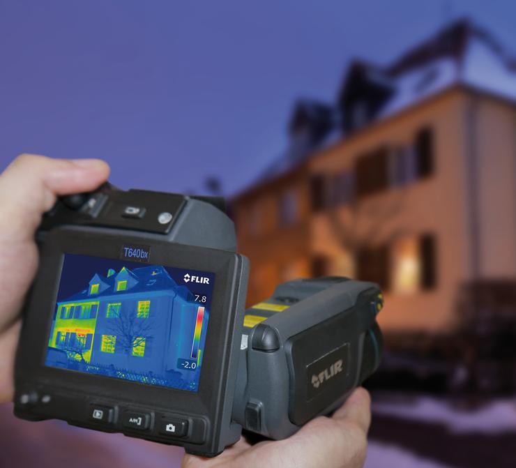 Haus mit schneebedecktem Dach wird mit einer Wärmebildkamera fotografiert, man sieht bunte Stellen auf de Bild für die Wärmezonen.